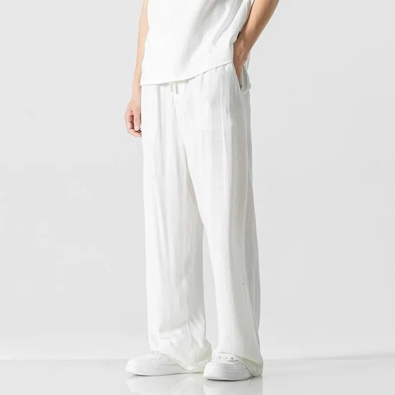 Men's beige loose straight-leg linen trousers with a drawstring waist, shown in a casual summer setting.