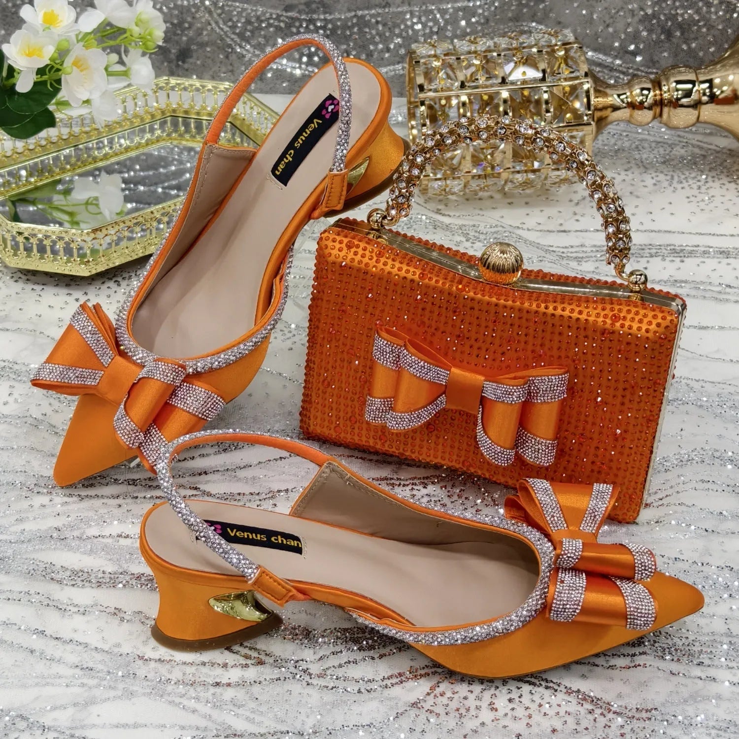 Women's black pointed sandals with a square rhinestone buckle and mid heel, next to a matching clutch bag.