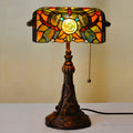 Tiffany-style desk lamp with coloured stained glass shade and resin base on a wooden desk.