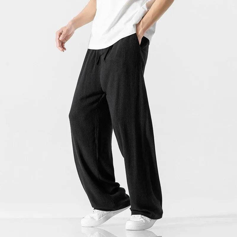 Men's beige loose straight-leg linen trousers with a drawstring waist, shown in a casual summer setting.