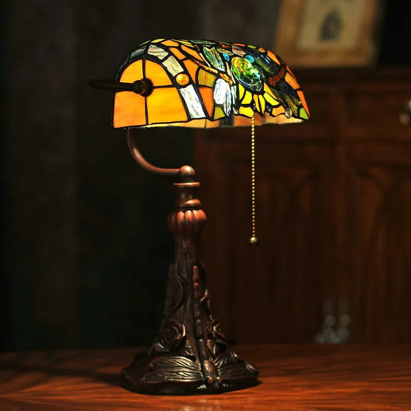 Tiffany-style desk lamp with coloured stained glass shade and resin base on a wooden desk.