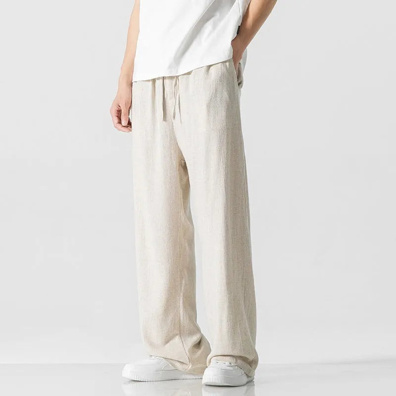 Men's beige loose straight-leg linen trousers with a drawstring waist, shown in a casual summer setting.