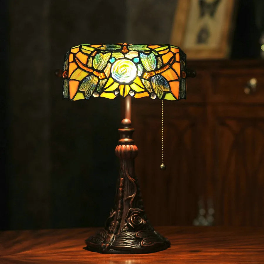 Tiffany-style desk lamp with coloured stained glass shade and resin base on a wooden desk.